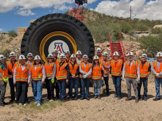 A group of people in white hard hats and orange vests stand in front of a large tire