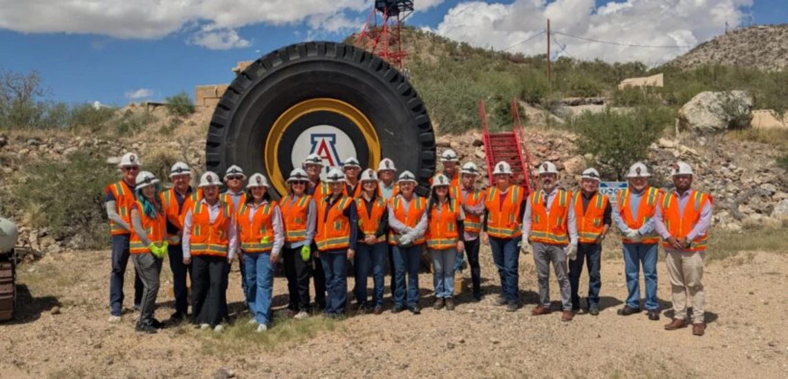 A group of people in white hard hats and orange vests stand in front of a large tire