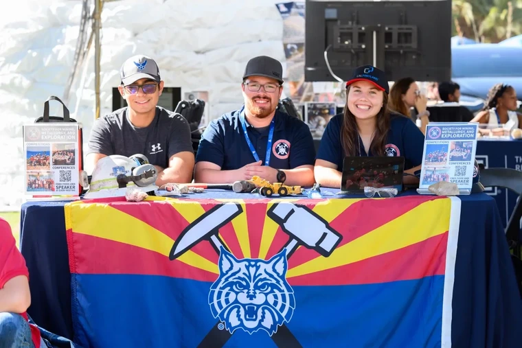 three people, seated at a table draped with a flag of Univ. of Arizona mascot and mining tools